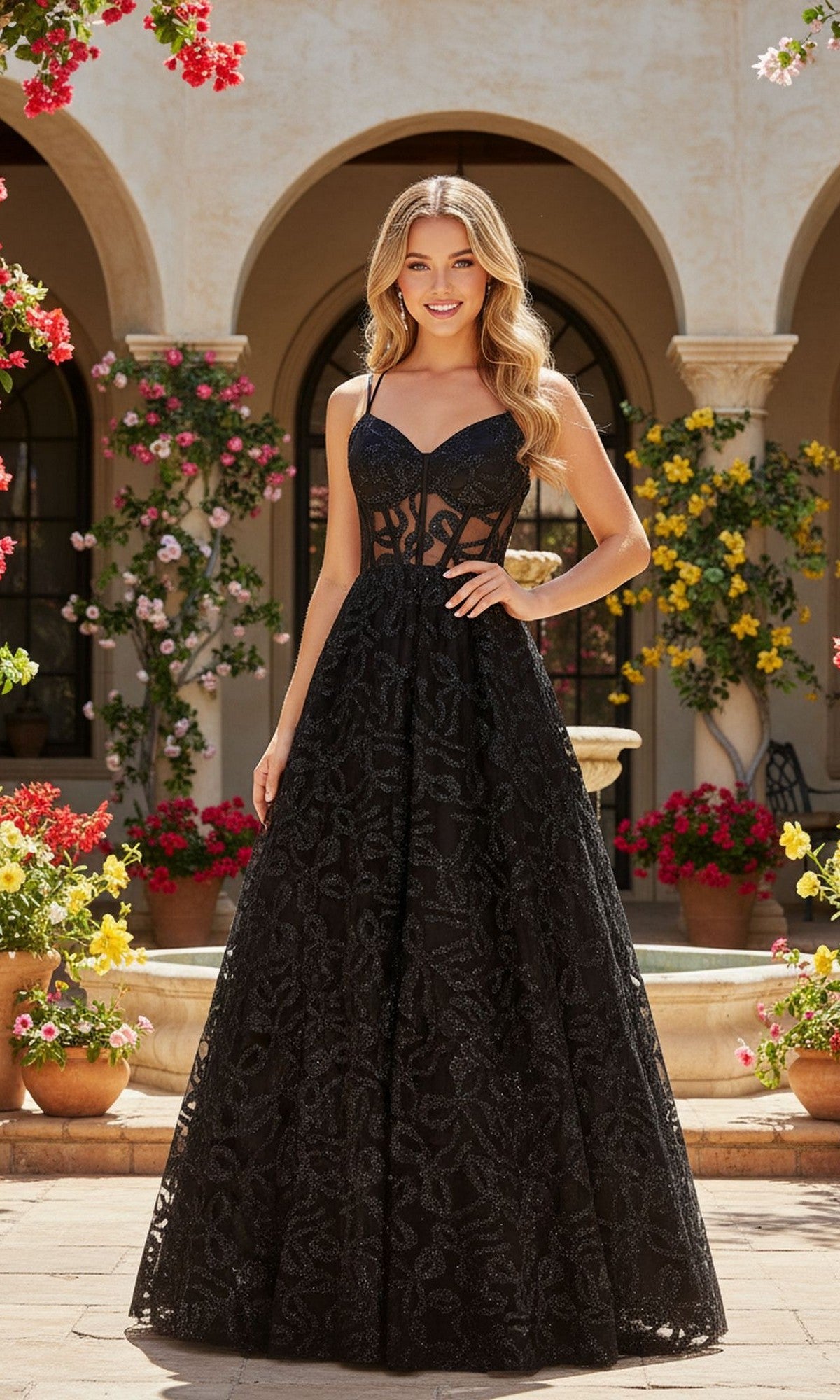 A woman wears the Long Prom Dress: Blondie Nites 6056BN in black, featuring a sleeveless lace design with a subtle corset bodice, as she stands outdoors in a garden courtyard with arched doorways and blooming flowers.